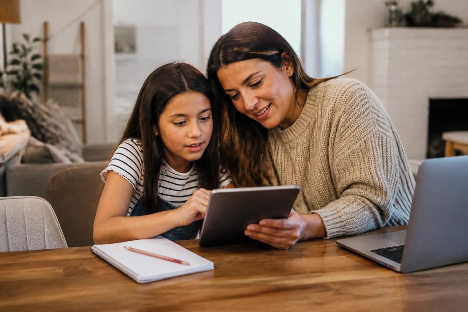 A parent and child looking at a tablet together at a dining table with a notebook beside it