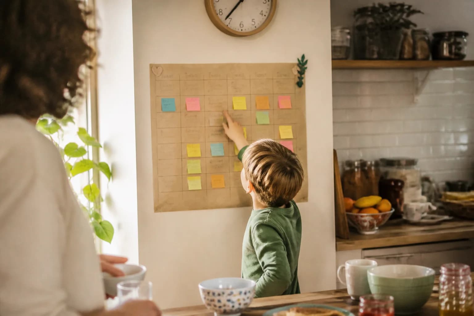 A large paper weekly schedule pinned to a kitchen wall with sticky notes, a child pointing at it