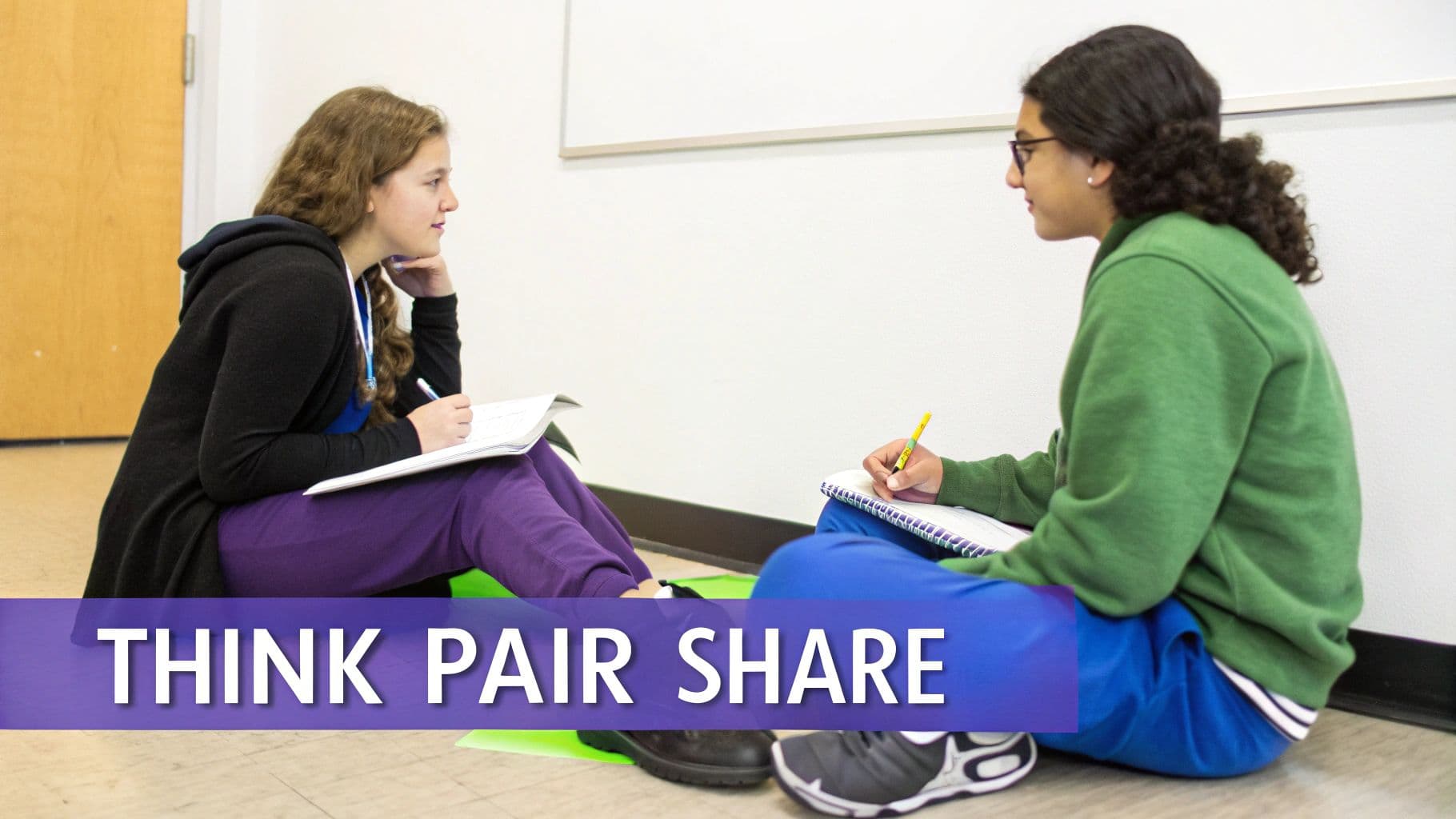 Two students sit on the floor, facing each other, writing in notebooks, embodying Think Pair Share.
