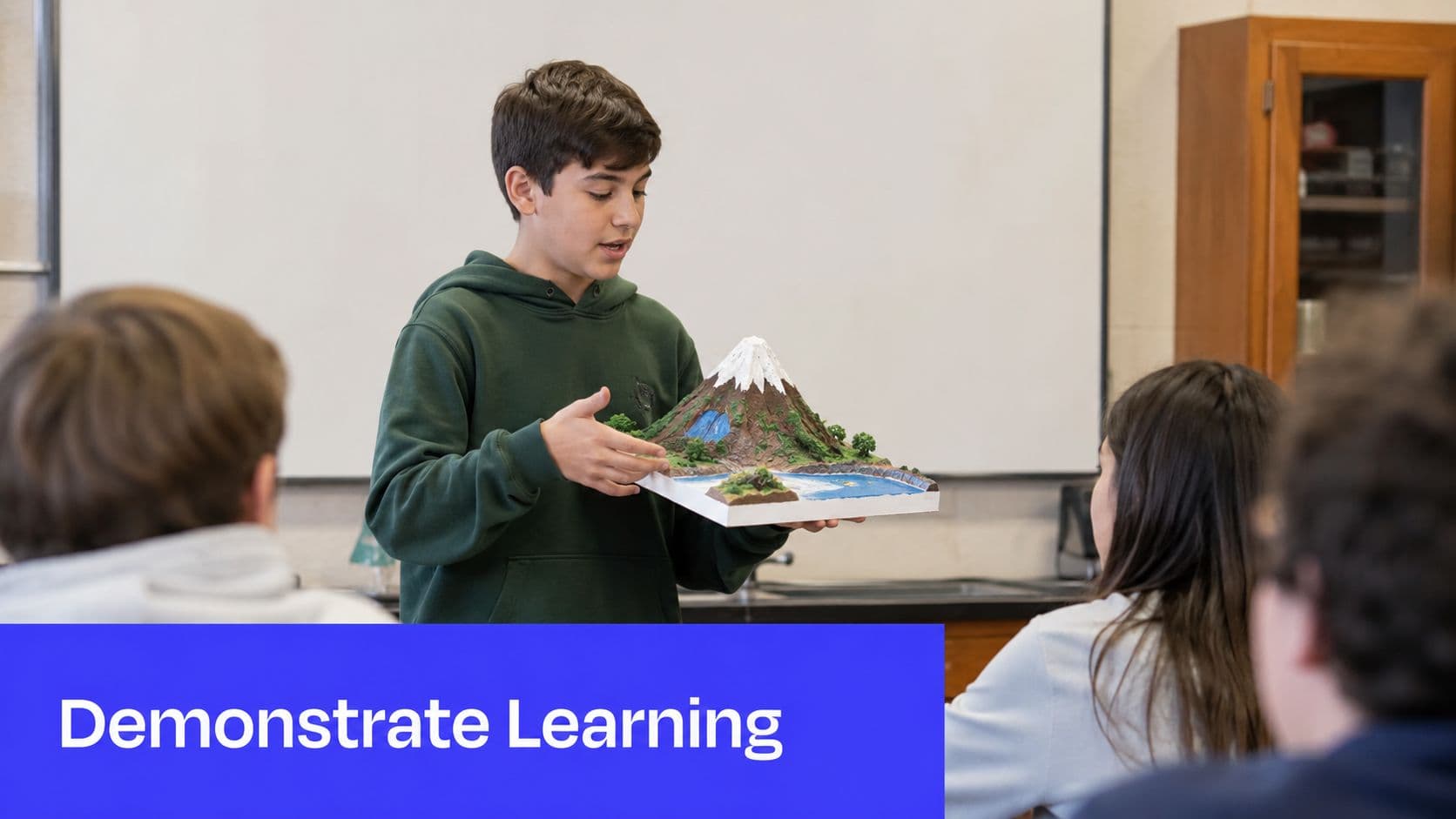 A male student standing in a classroom presenting a diorama of a mountain to his classmates.