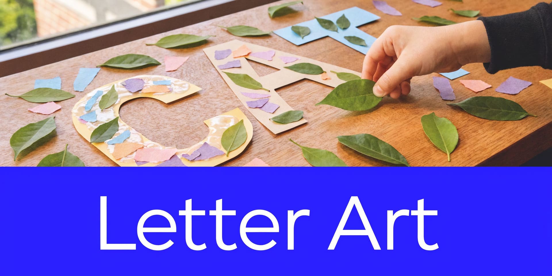 A child's hand decorating cardboard letters with green leaves and colorful paper pieces on a wooden table.