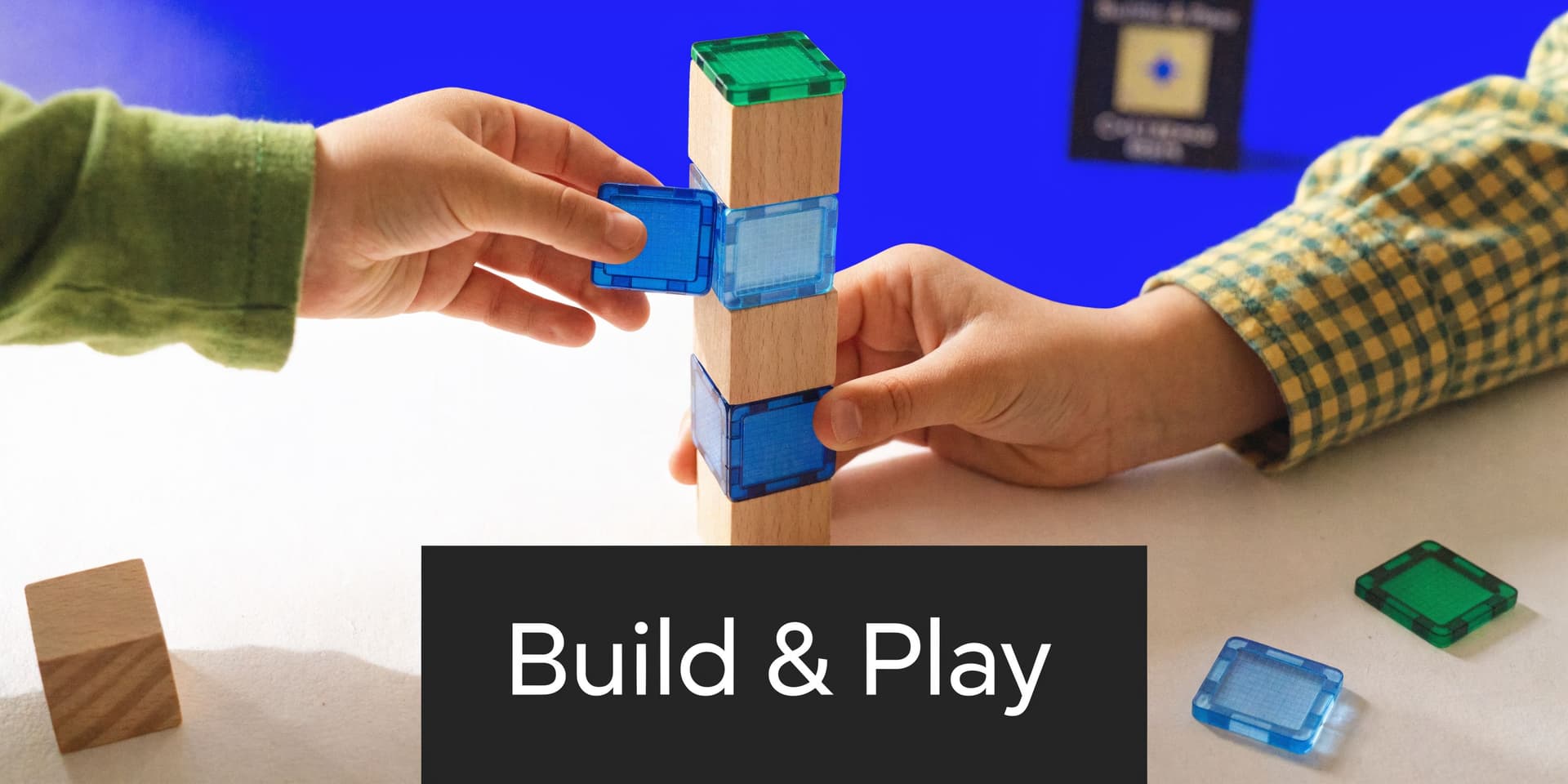 Two children's hands interacting to build a tower using wooden blocks and magnetic colorful plastic tiles.