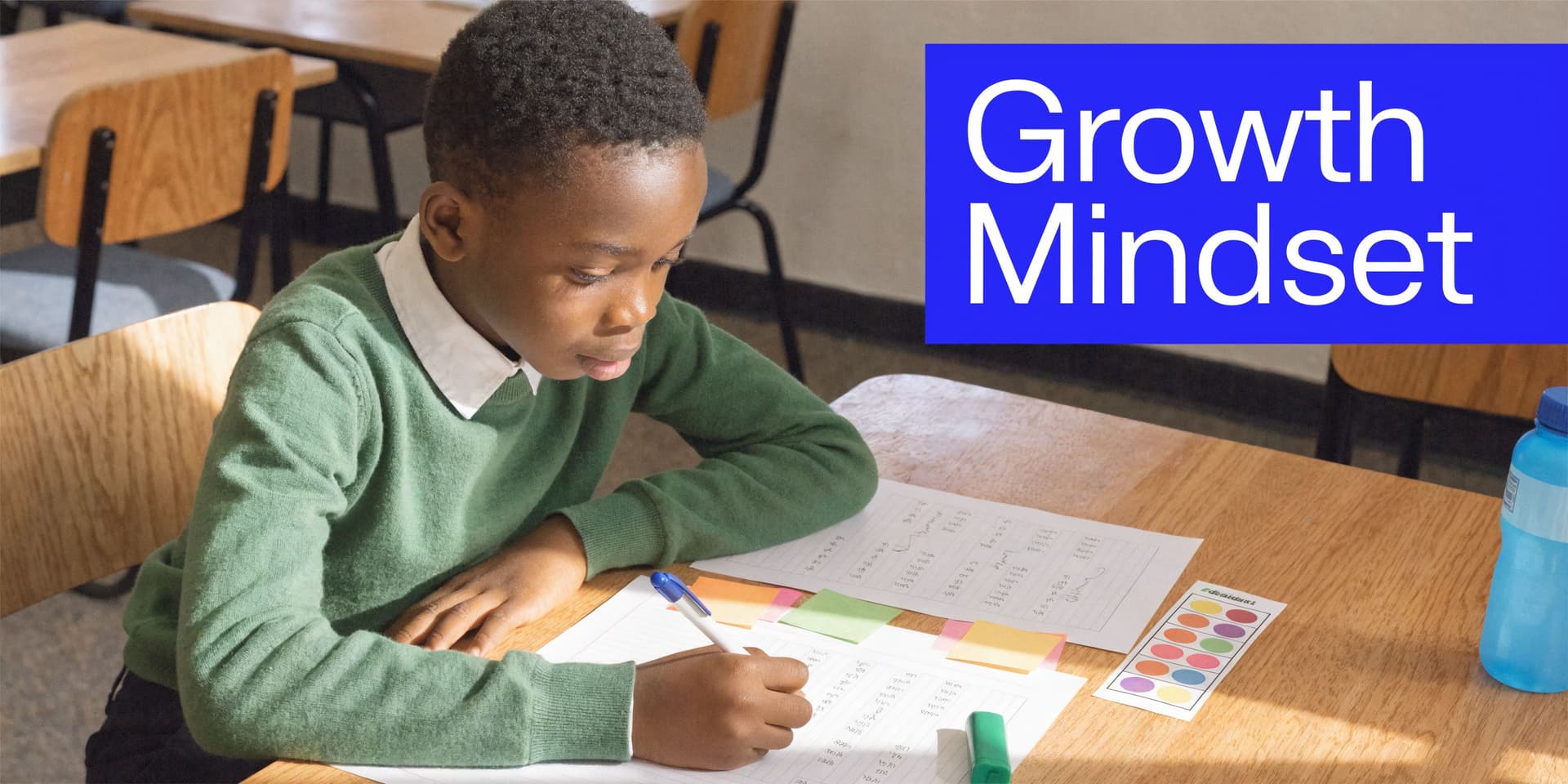 A young schoolboy in a green sweater focuses intently on writing in his workbook at a desk.