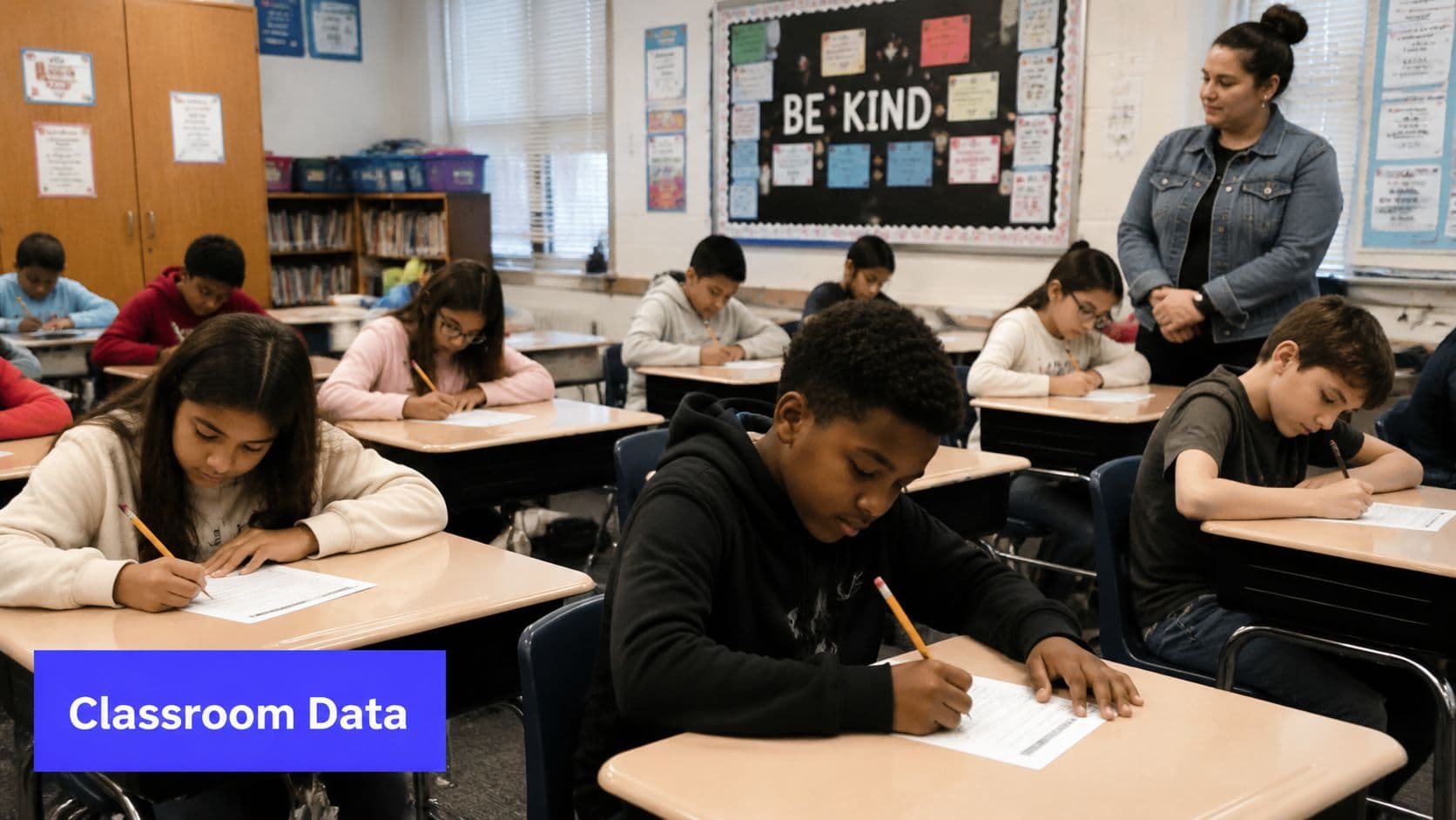 A teacher stands in a classroom while students concentrate on their desk work and data collection tasks.